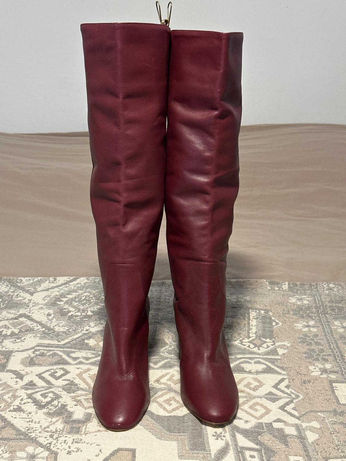 Burgundy leather knee-high boots on a rug with a geometric design in beige and grey tones. Neutral background.
