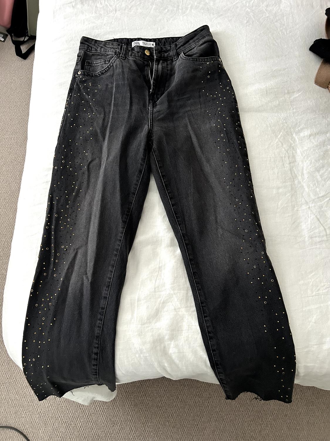 Black denim jeans with gold studs on the legs, laid on a white bed.