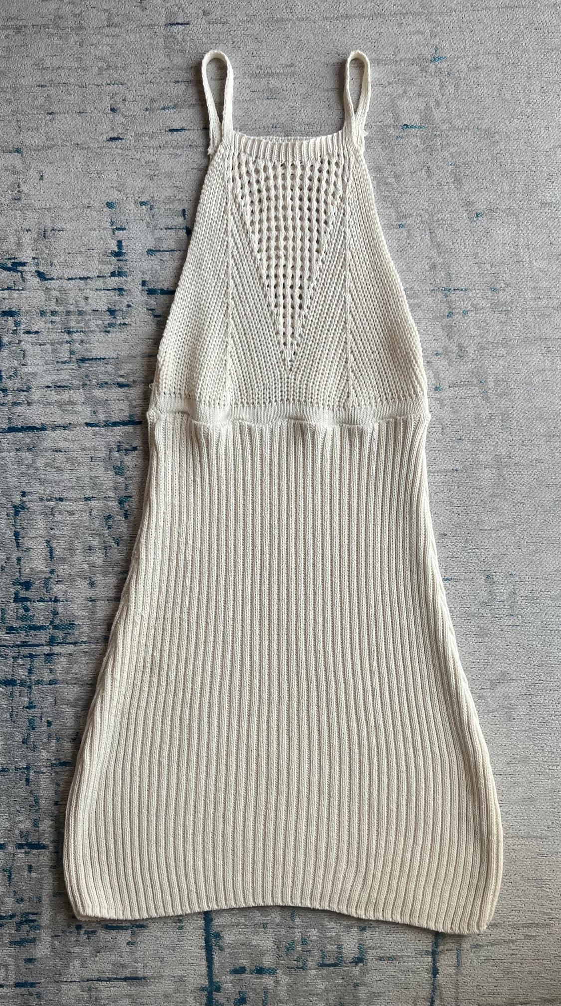 Beige knitted dress with thin straps and an openwork knit design on the upper part, against a textured grey background.