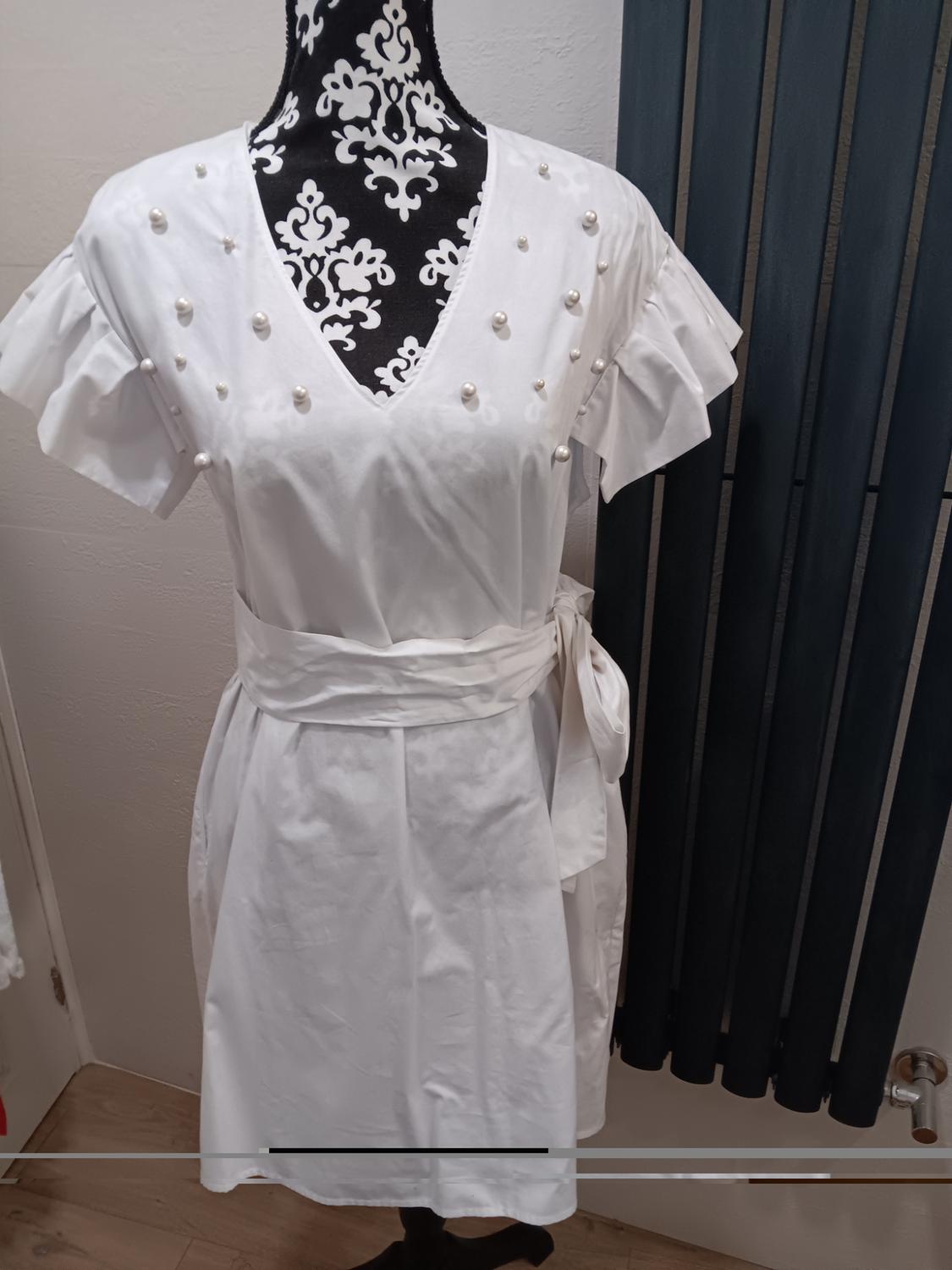 Mannequin with a short-sleeved white dress with pearls on the bust and a belt, against a white wall and dark curtain background.