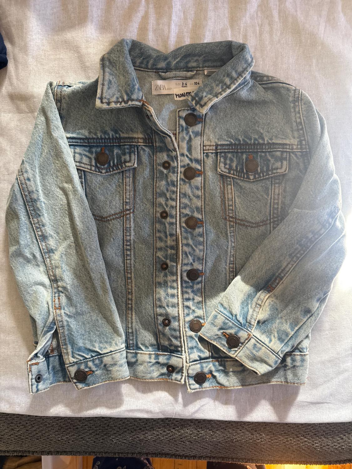 Light blue denim baby jacket with brown buttons and front pockets, on a white surface. Visible label.