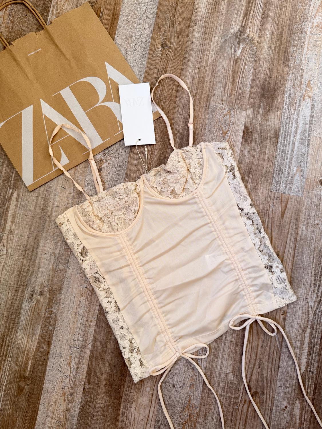 Beige camisole top with lace and bows on a wooden floor, next to a paper bag bearing the text ZARA.