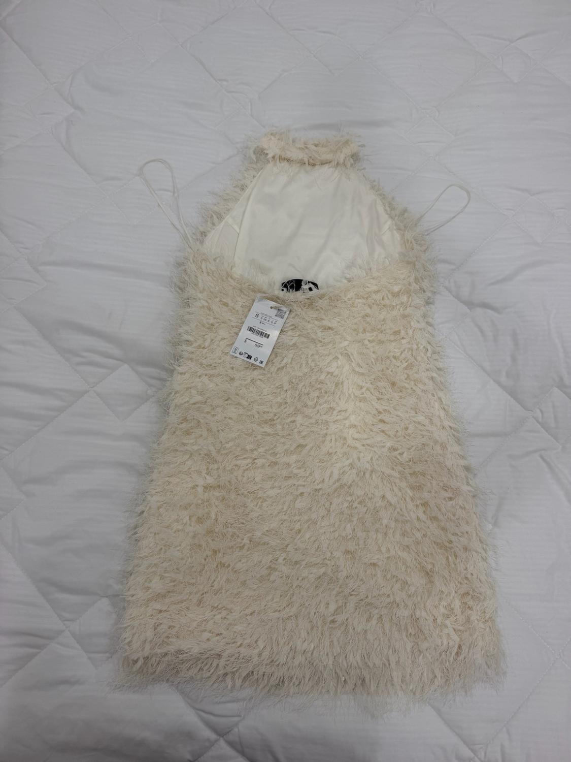 Cream halter-neck mini dress with a furry texture, flat lay on a white quilted duvet. Shows a tag.