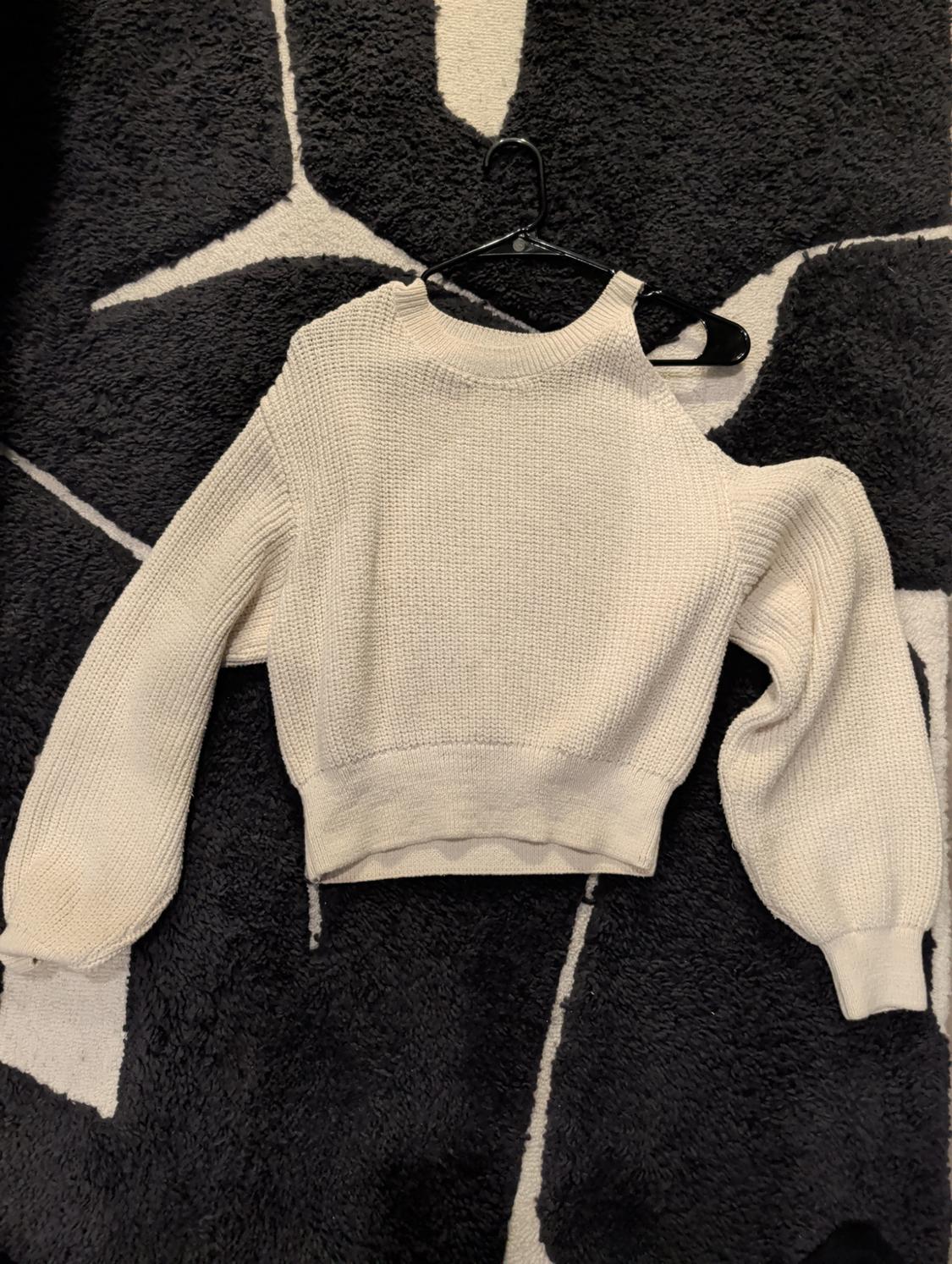 Chunky knit beige jumper with one exposed shoulder, hanging on a black hanger over a black and white geometric patterned rug.