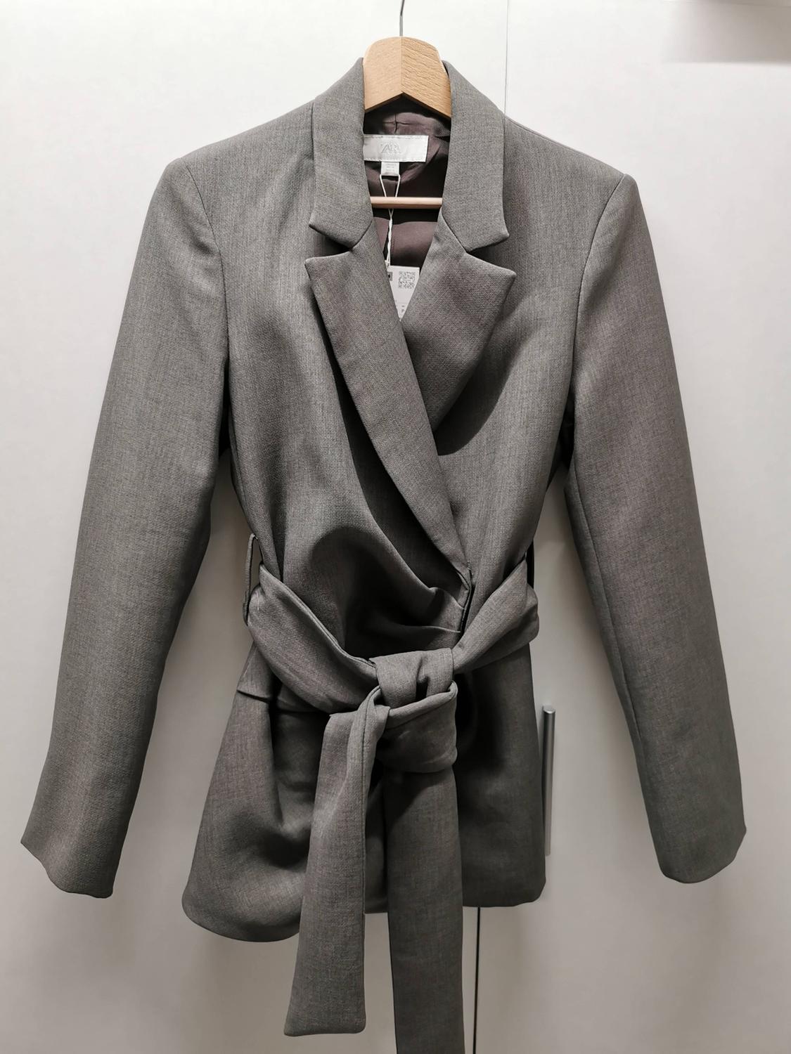 Grey jacket with a tie belt, hanging on a wooden hanger against a white background. Visible label on the inside.