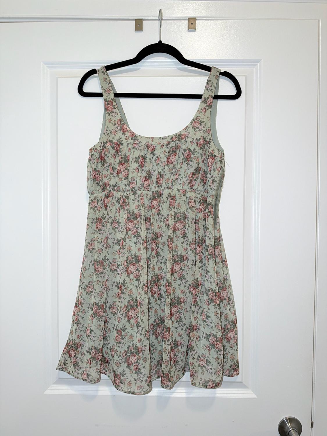 Short sleeveless dress with a pink floral print on a light green background, hanging on a black hanger on a white door.