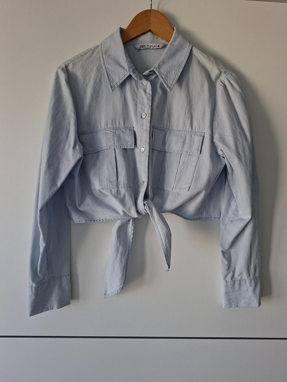 Light blue long-sleeved shirt hanging on a wooden hanger against a white background. It has front pockets, a knot at the hem and a ZARA label.