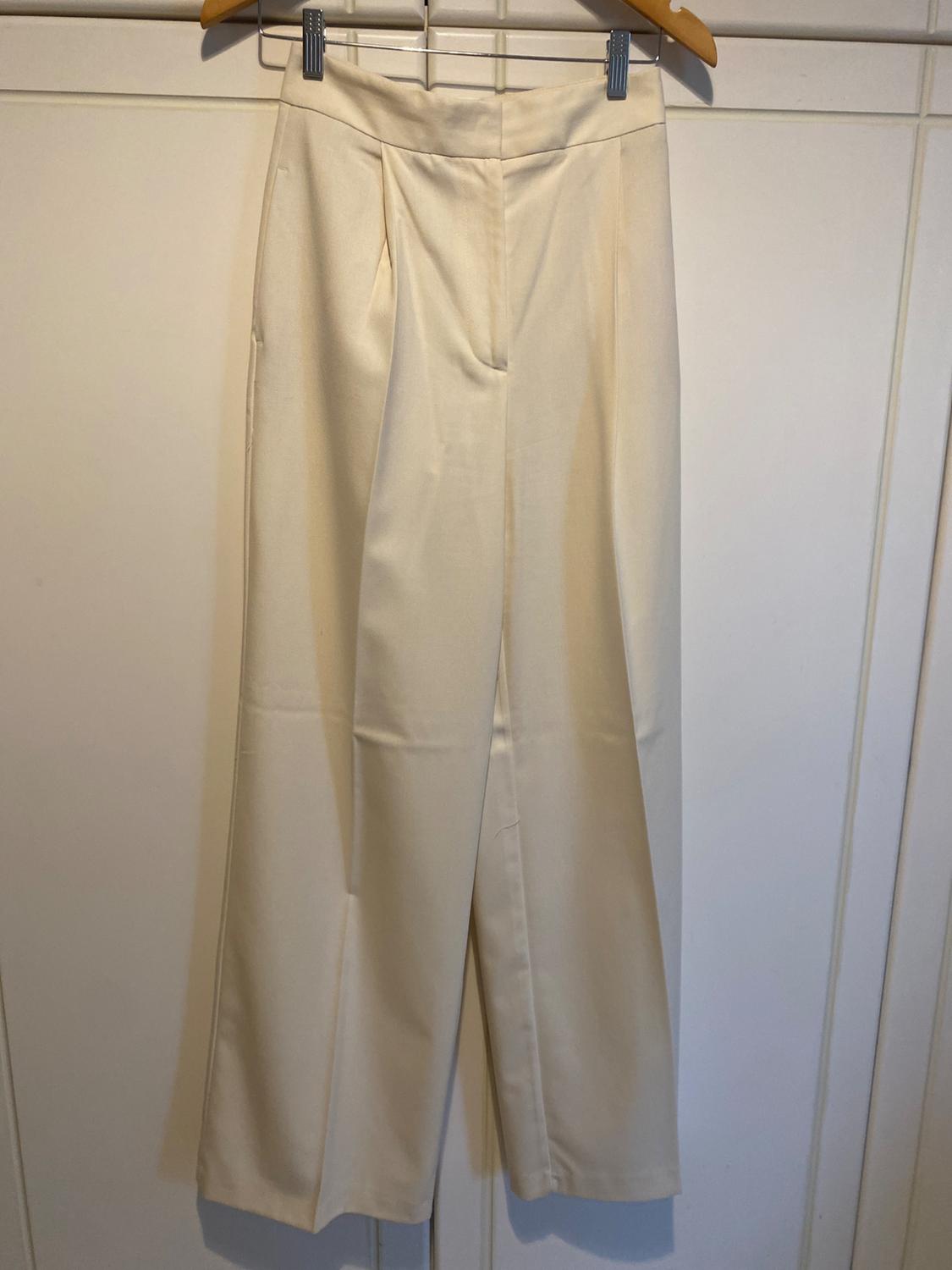 Cream-coloured wide-leg trousers with pleats, hanging on a wooden hanger in front of a white wardrobe. Minimalist and relaxed style.