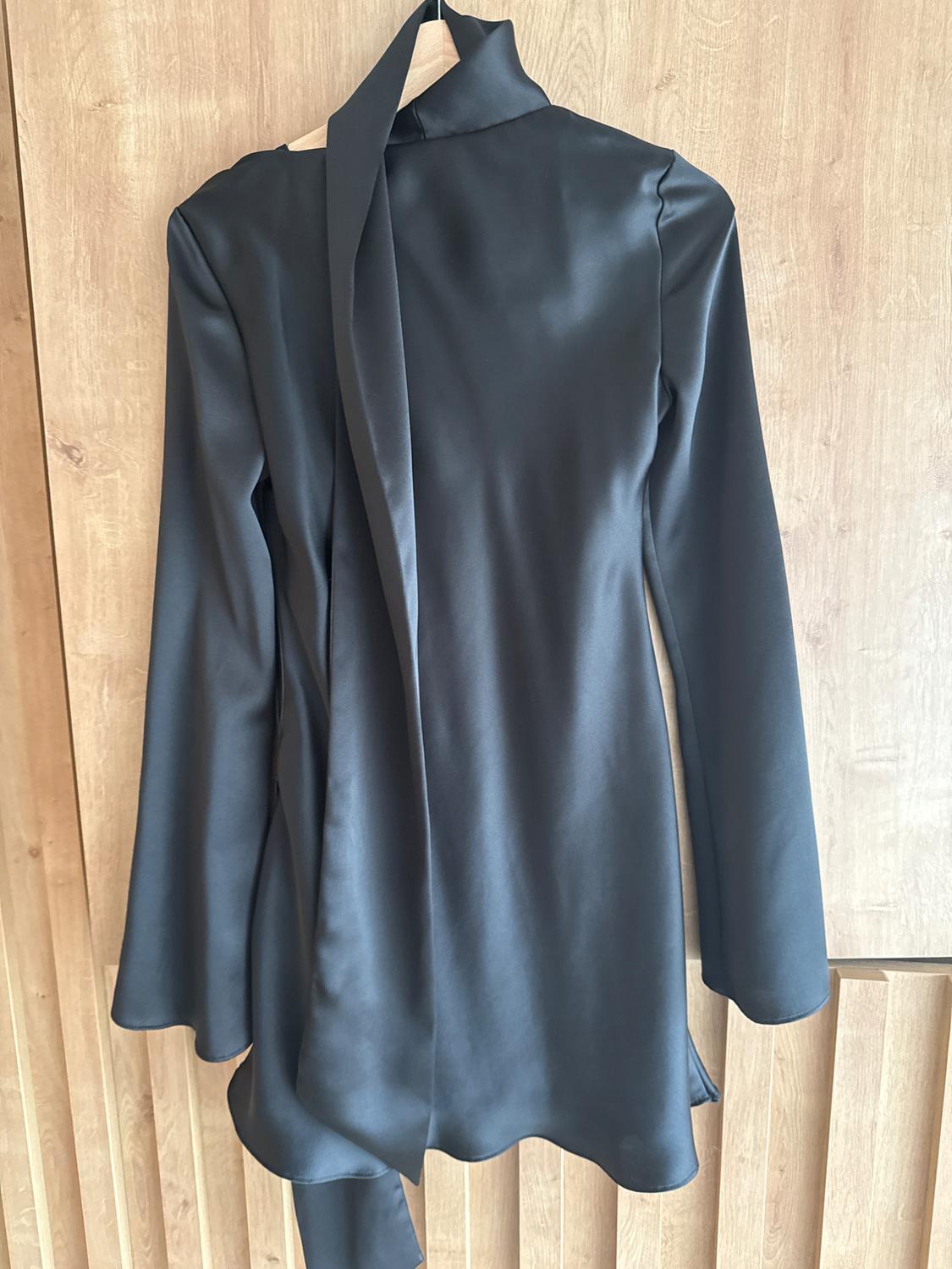 Black satin high-neck bow dress, hanging on a wooden hanger against a light wooden background.
