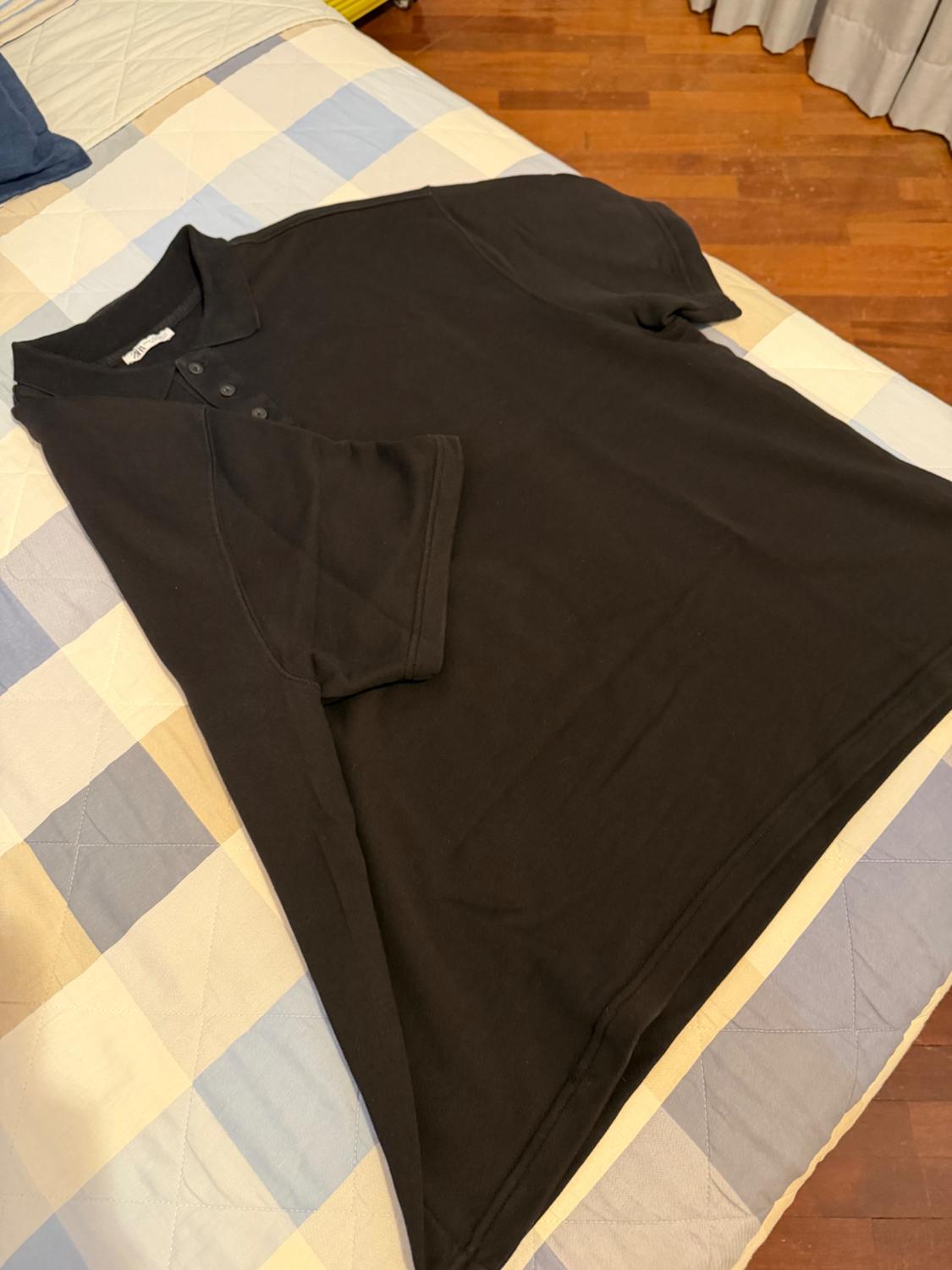 Black short-sleeved polo shirt on a bed with a blue and beige checked bedspread, in a room with a wooden floor.