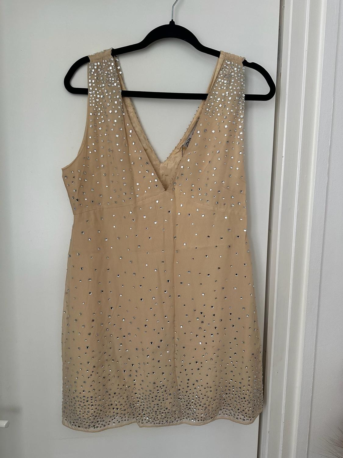 Beige short dress with a V-neck and silver appliqués, hanging from a black hanger on a white door. It reads ZARA.