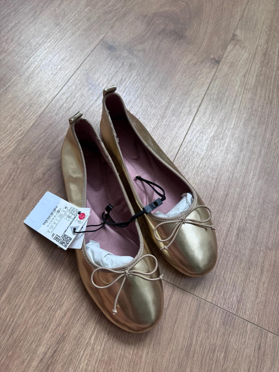 Pair of gold ballet flats with a front bow and label, placed on a light wooden floor.