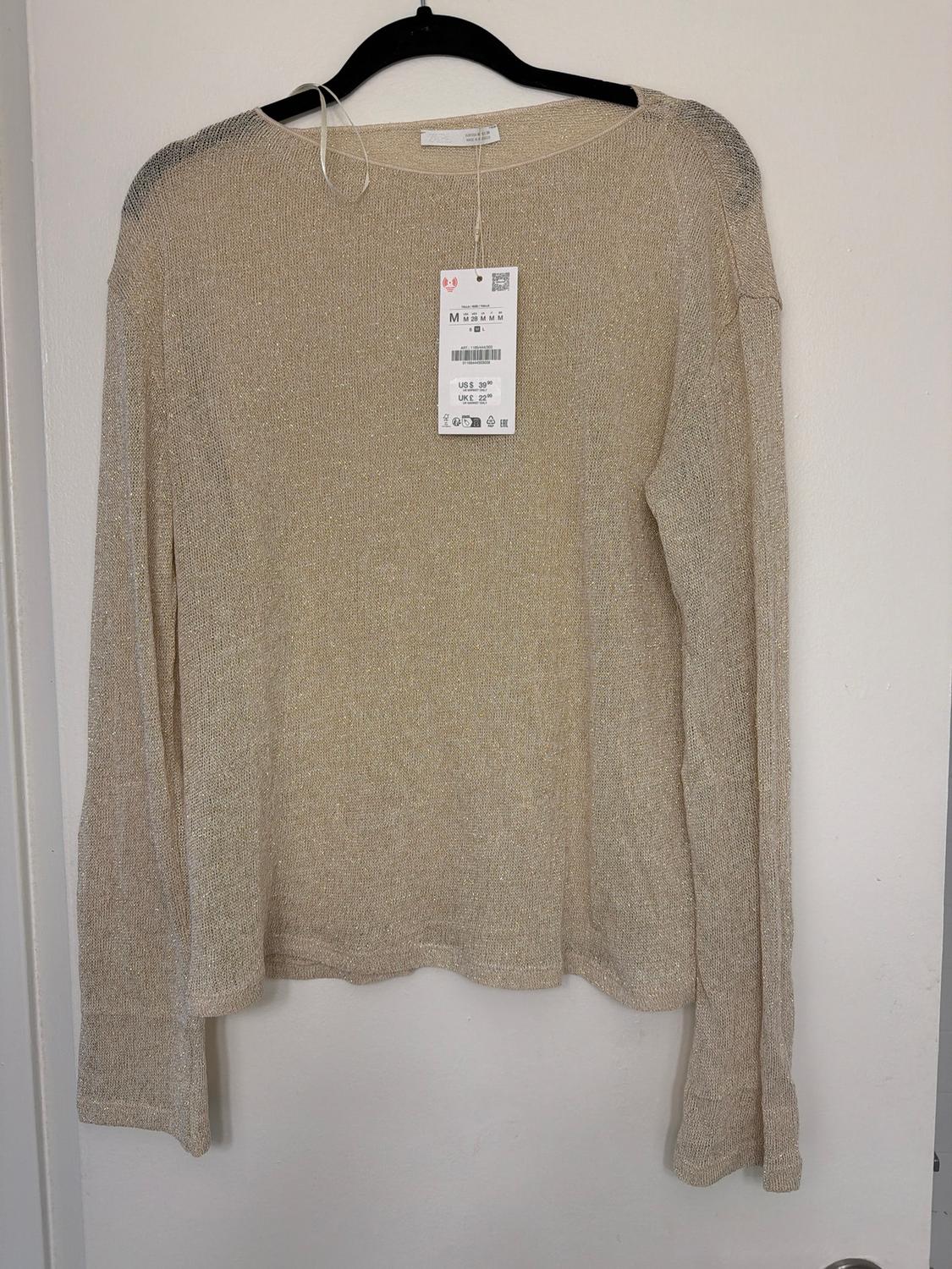 Beige long-sleeved jumper hanging on a black hanger, with visible price tag showing size M and price US$ 29.95.