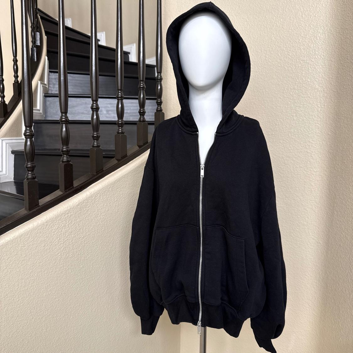 Mannequin wearing a black zip-up hoodie. Casual indoor setting next to a dark staircase and a beige wall.