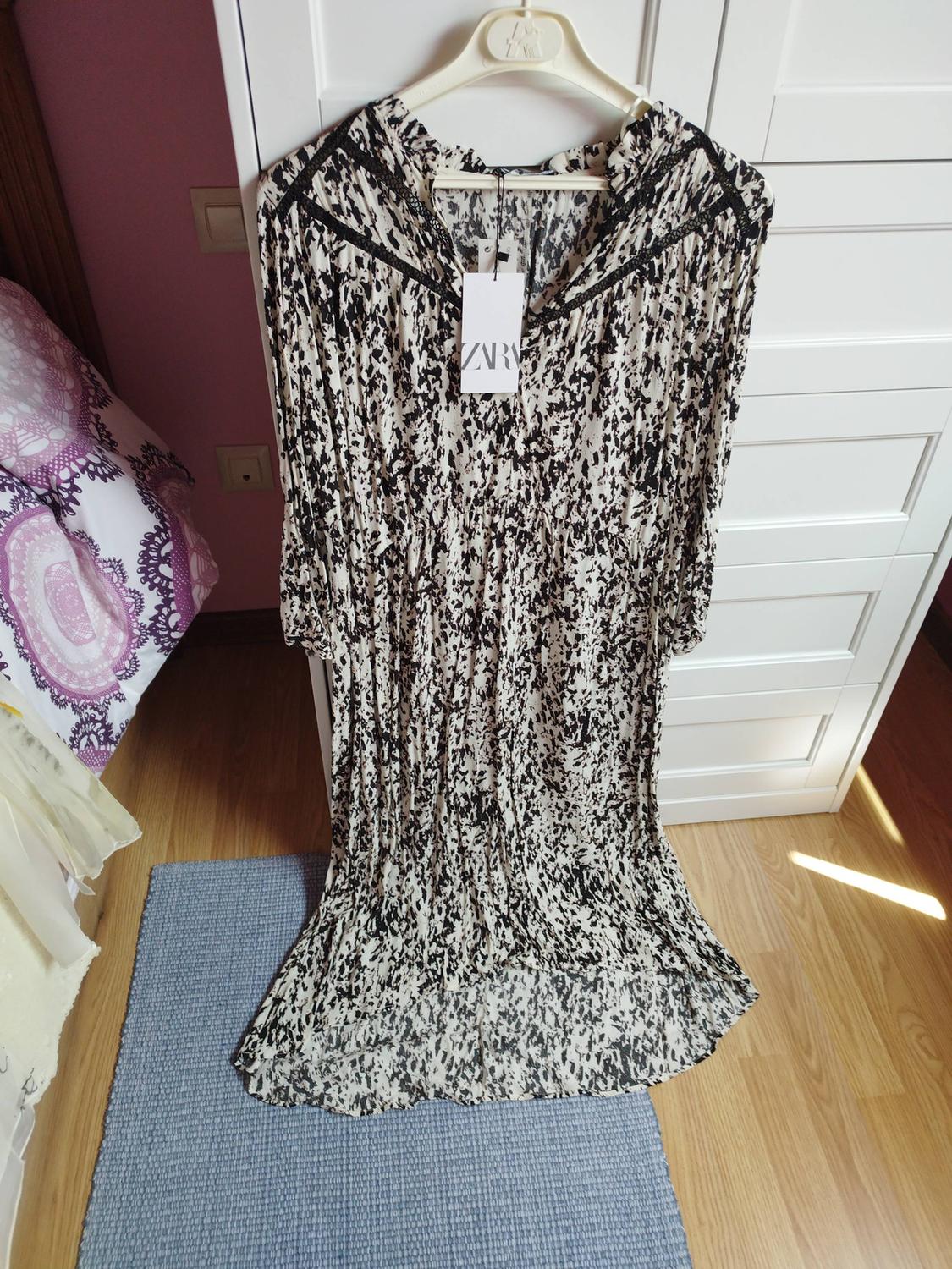 Black and white patterned dress hanging on a hanger above a blue rug. Label with "ZARA" text visible.
