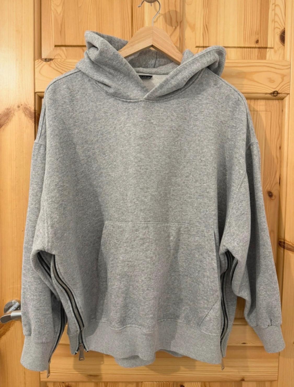 Grey hooded sweatshirt hanging on a coat hanger on a wooden door. It has a kangaroo pocket and zips on the side seams.