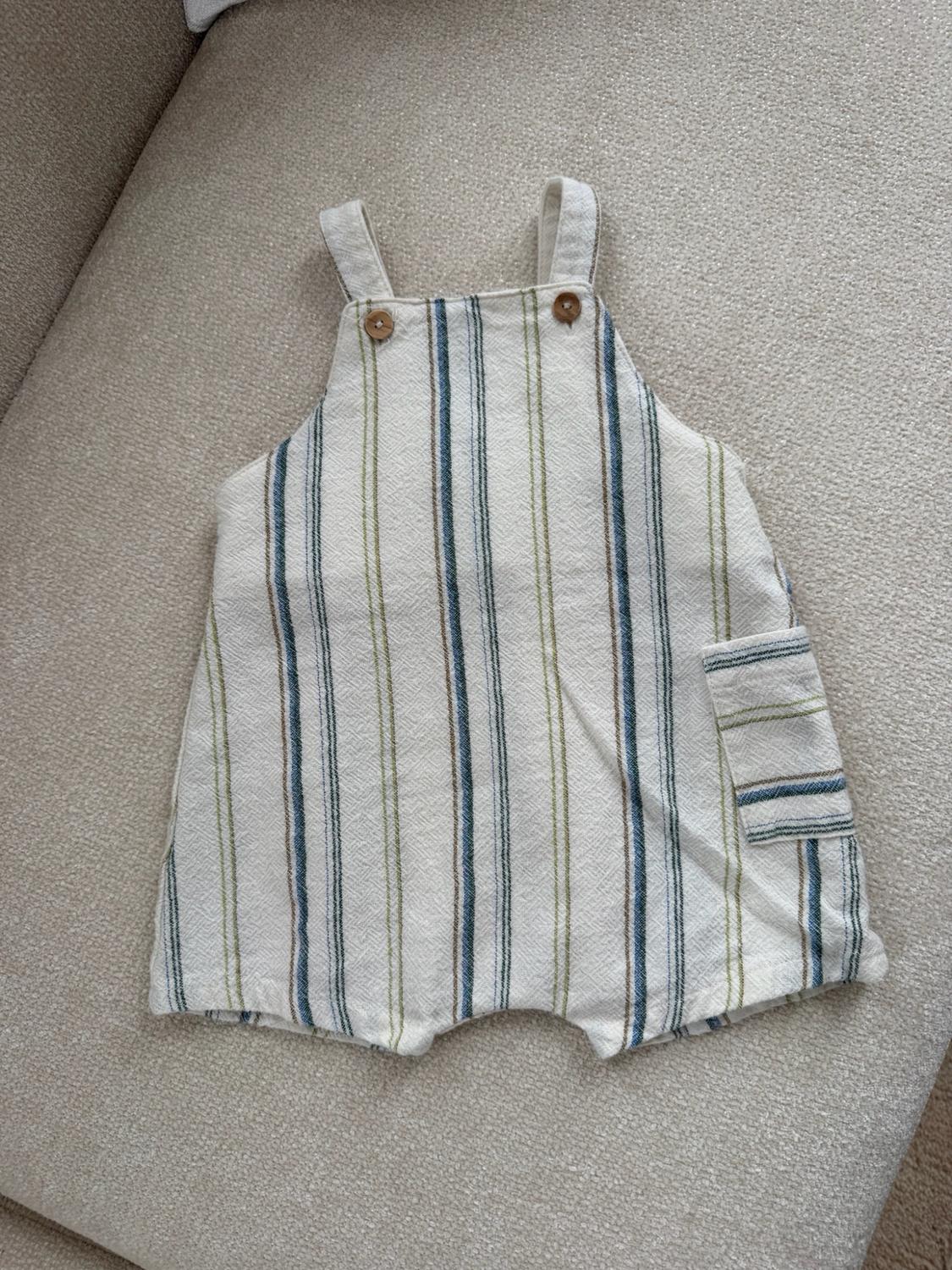 Cream-colored baby blanket with vertical stripes in blue, green, and brown. It features two wooden buttons on the straps and a side pocket.