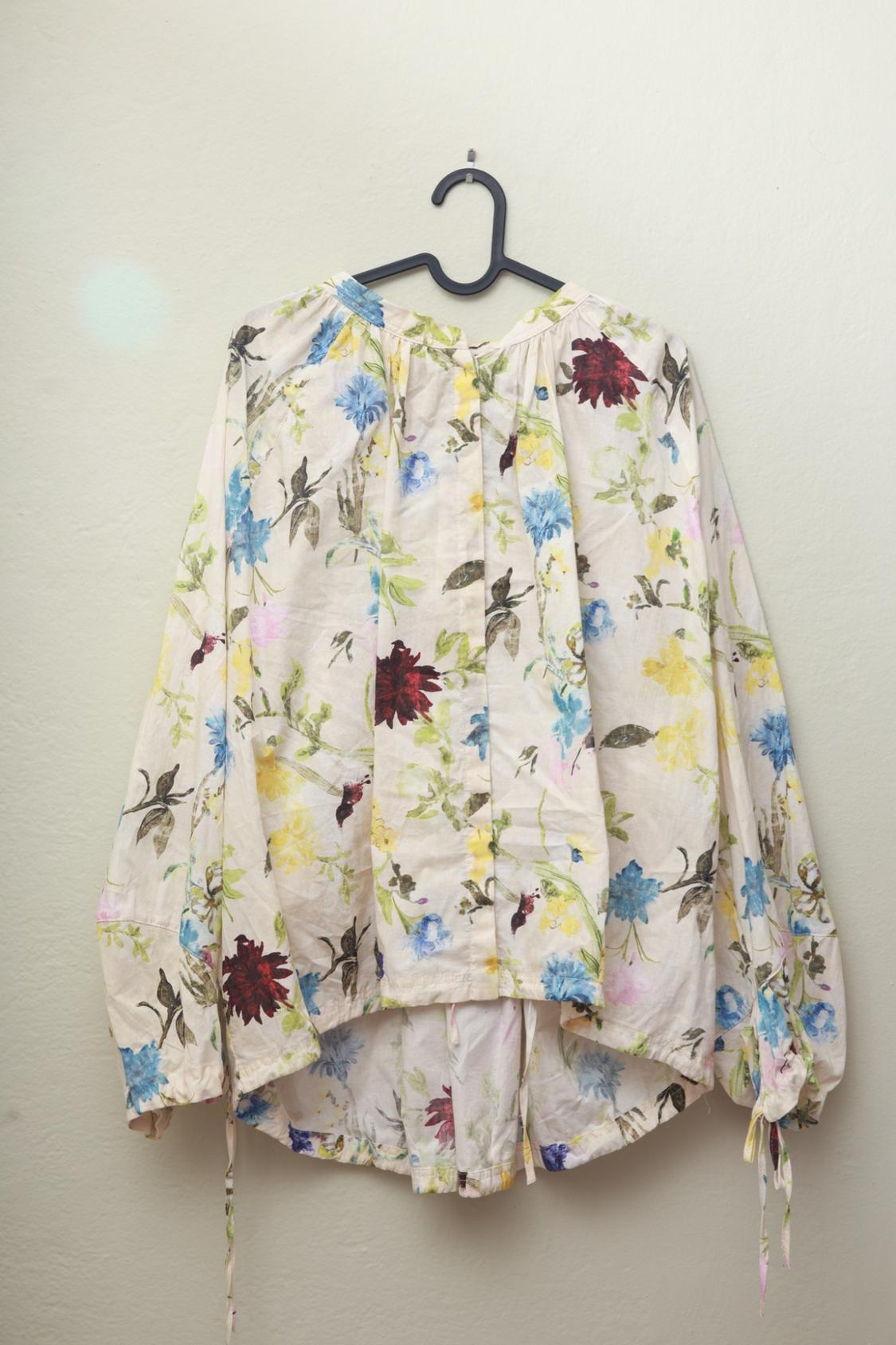Loose-fitting, long-sleeved floral print blouse with tie cuffs, hanging on a black hanger against a light-colored wall.