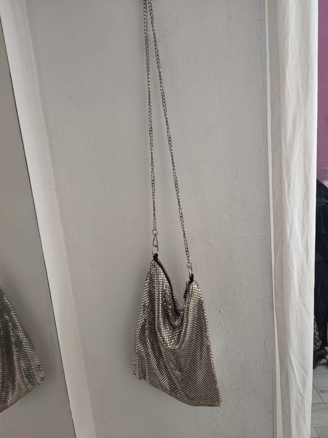 Metal mesh bag hanging on the white wall, reflected in a mirror. Background with white curtain and lilac wall.