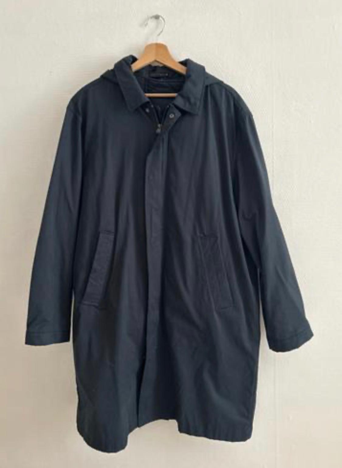 Dark long coat hanging on a wooden hanger against a light-coloured wall. Minimalist and relaxed composition focused on the garment.