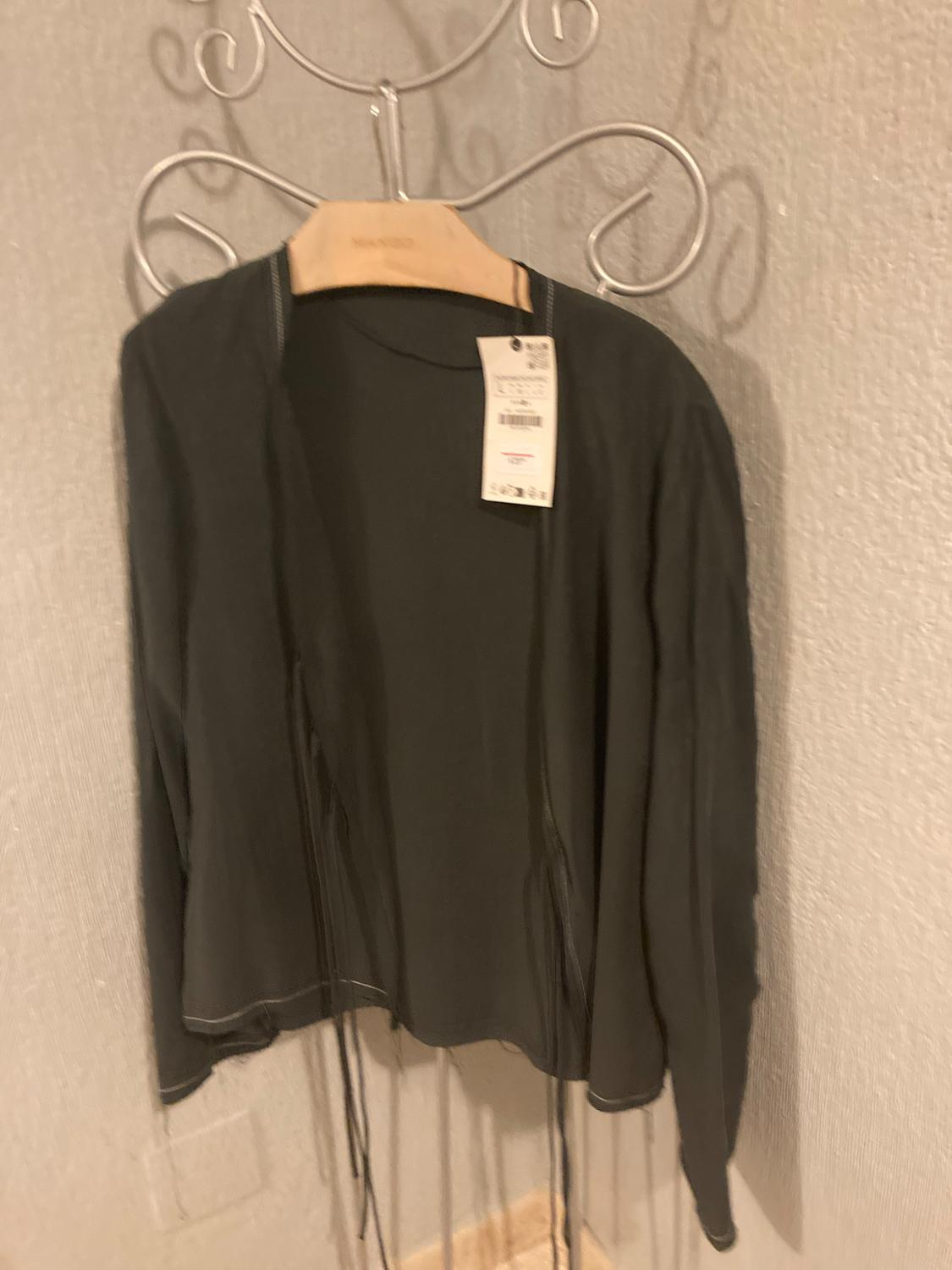 Dark grey top with label and hanging straps, on a wooden hanger with MANGO text, on a metal clothes rail in front of a light-coloured wall.
