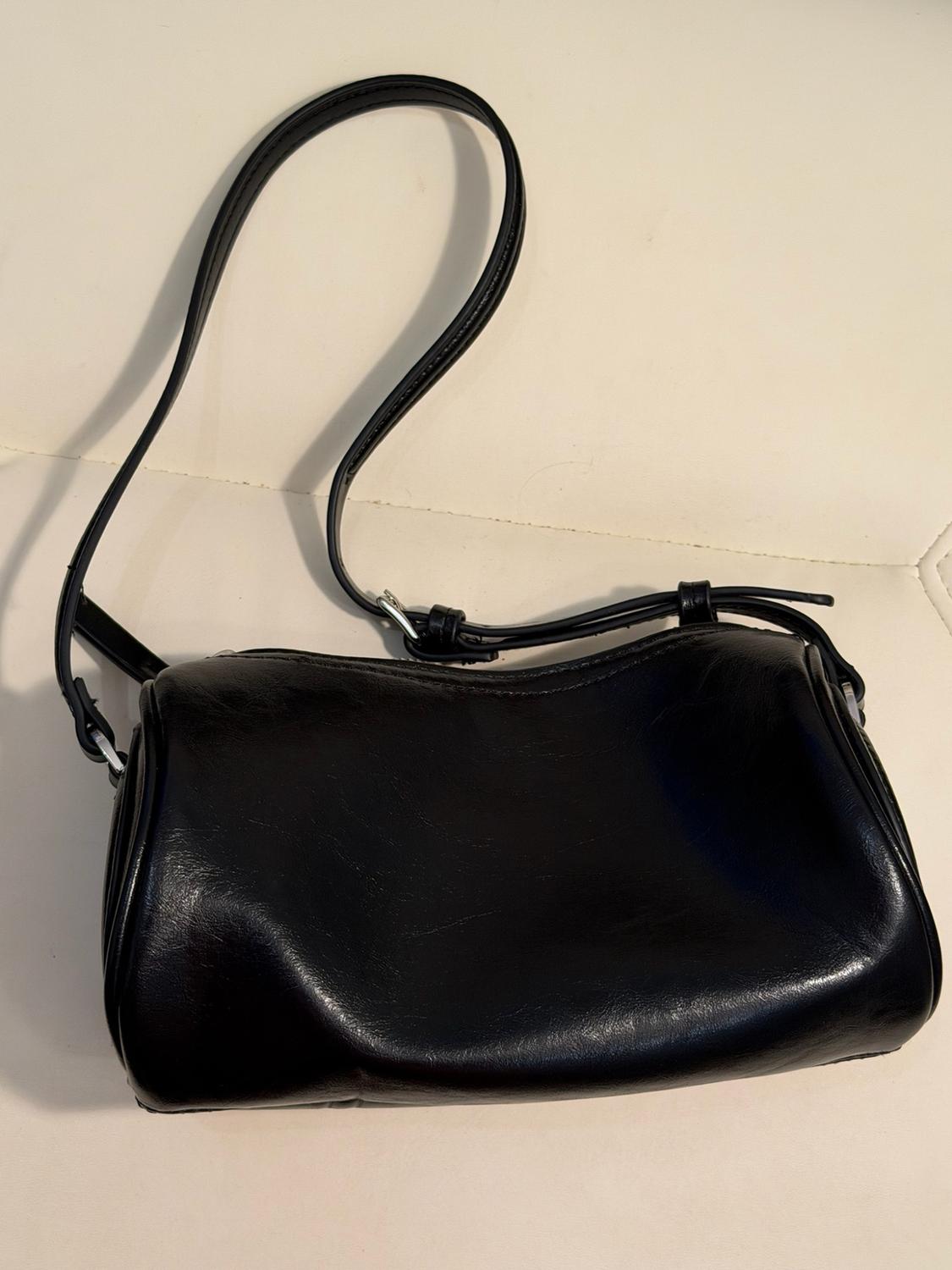 Black glossy handbag with a long adjustable strap and silver hardware, resting on a cream-coloured padded surface.