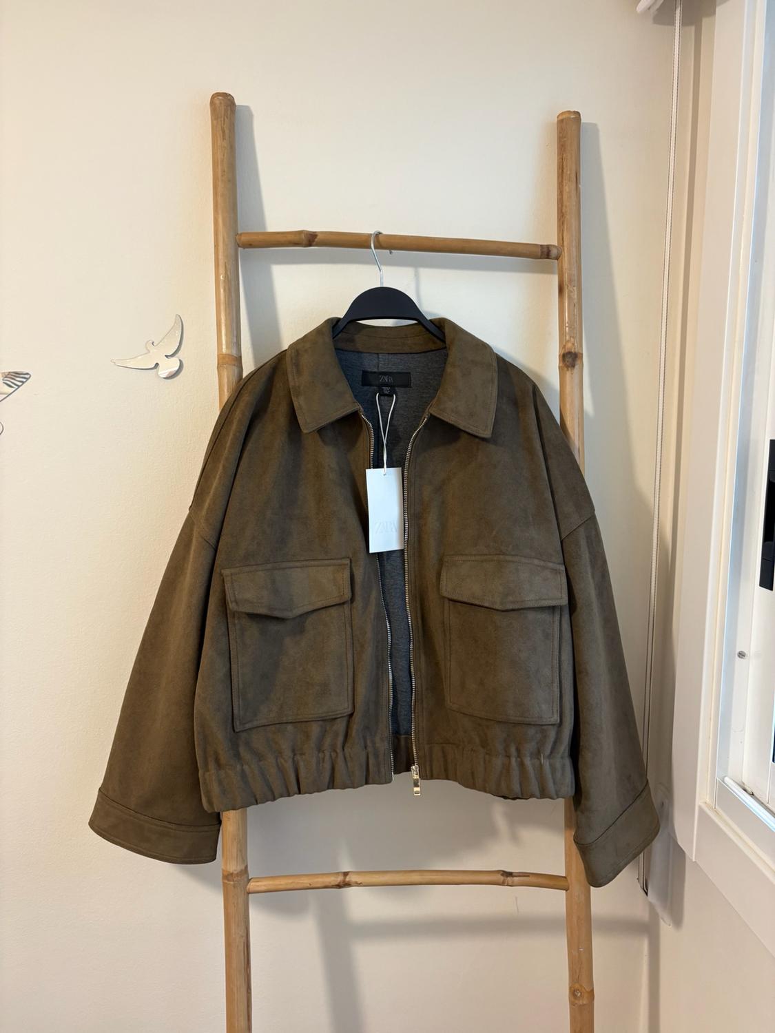 Brown jacket with a zip and front pockets hanging on a wooden ladder leaning against a white wall. ZARA label.
