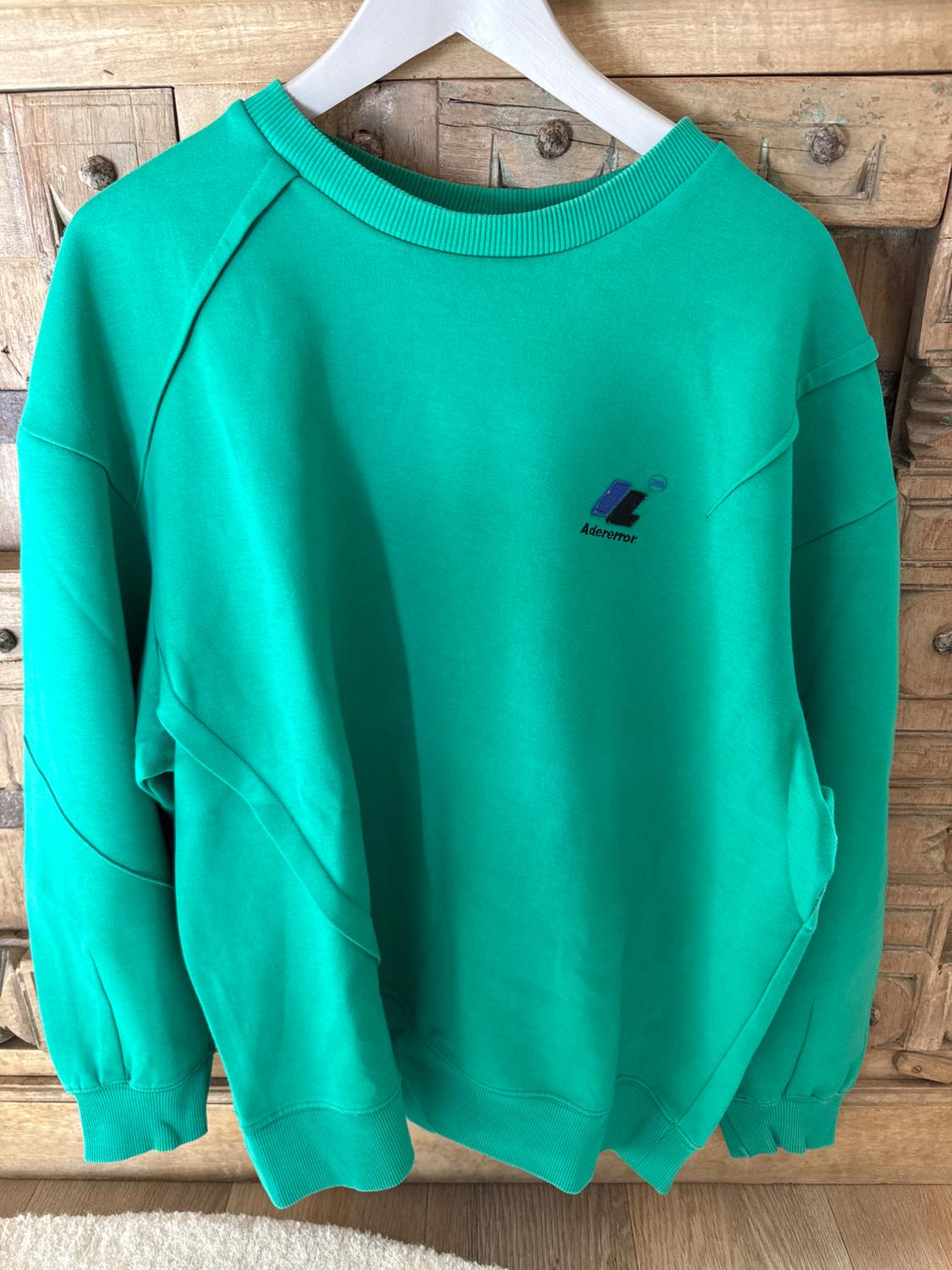 Green sweatshirt hanging on a white hanger in front of a wooden cabinet. "Adererror" logo on the chest.
