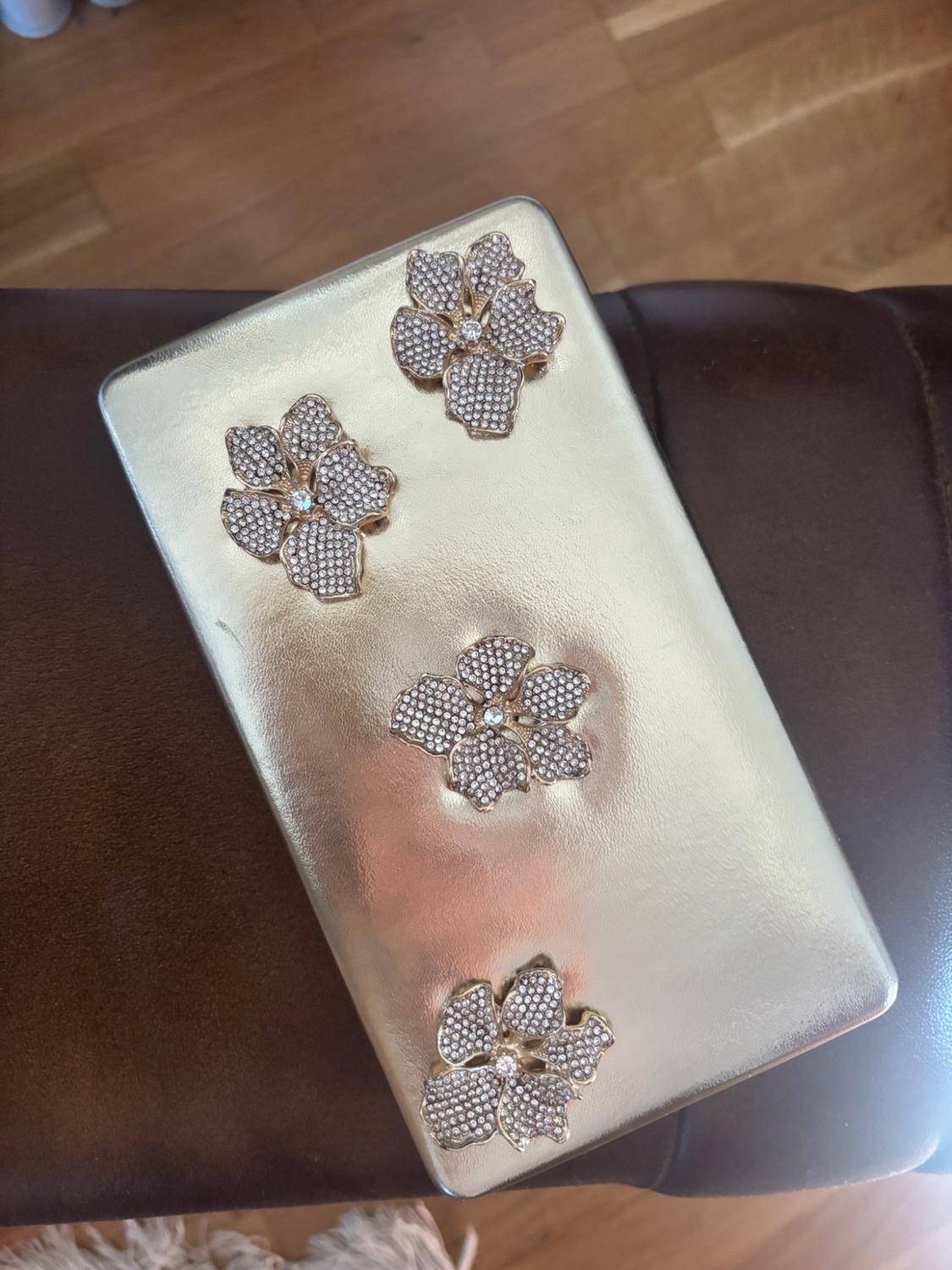 Rigid gold clutch bag with four floral appliqués made of sparkling gemstones, resting on a brown leather seat.