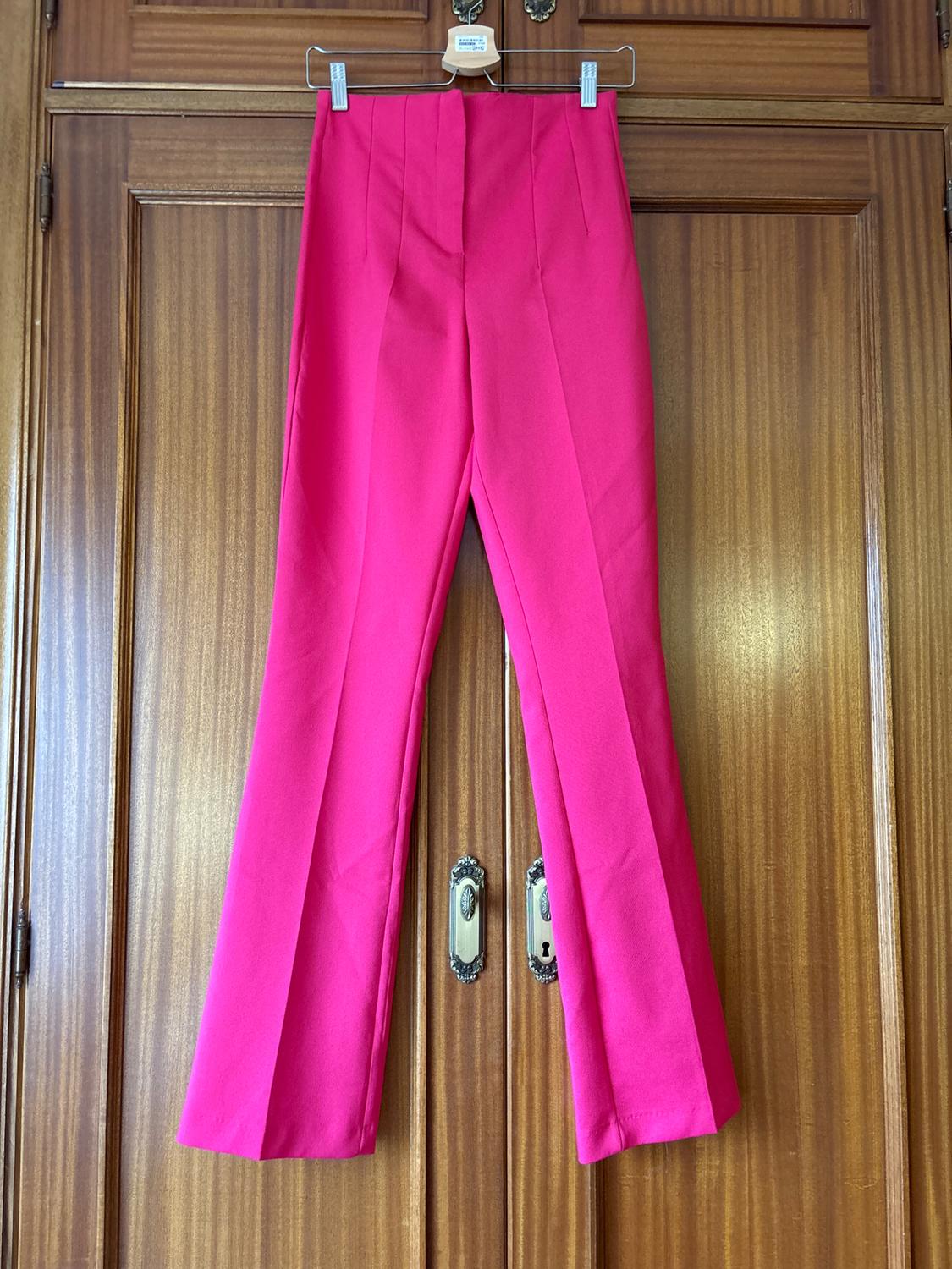 Fuchsia trousers hanging on a hanger over a wooden door. The garment has a wide-leg design and front pleats.