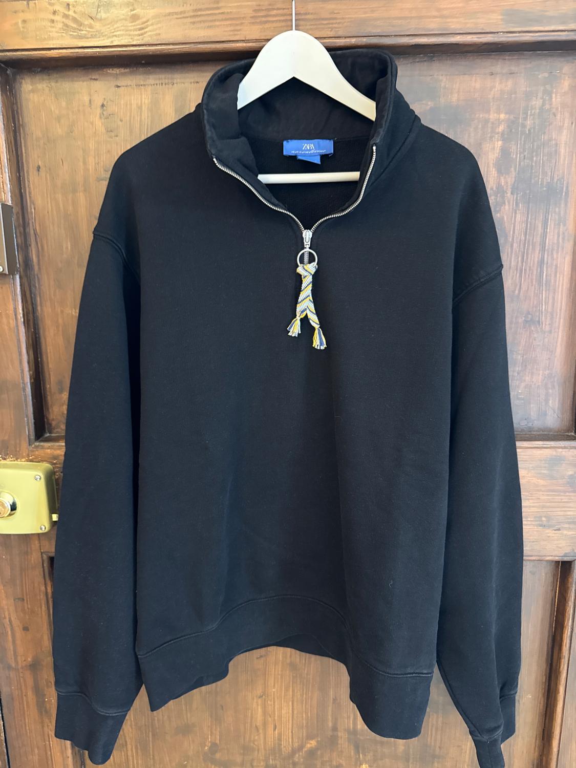 Black sweatshirt with a zip and decorative drawstring at the neck, hanging on a white hanger in front of a wooden door.