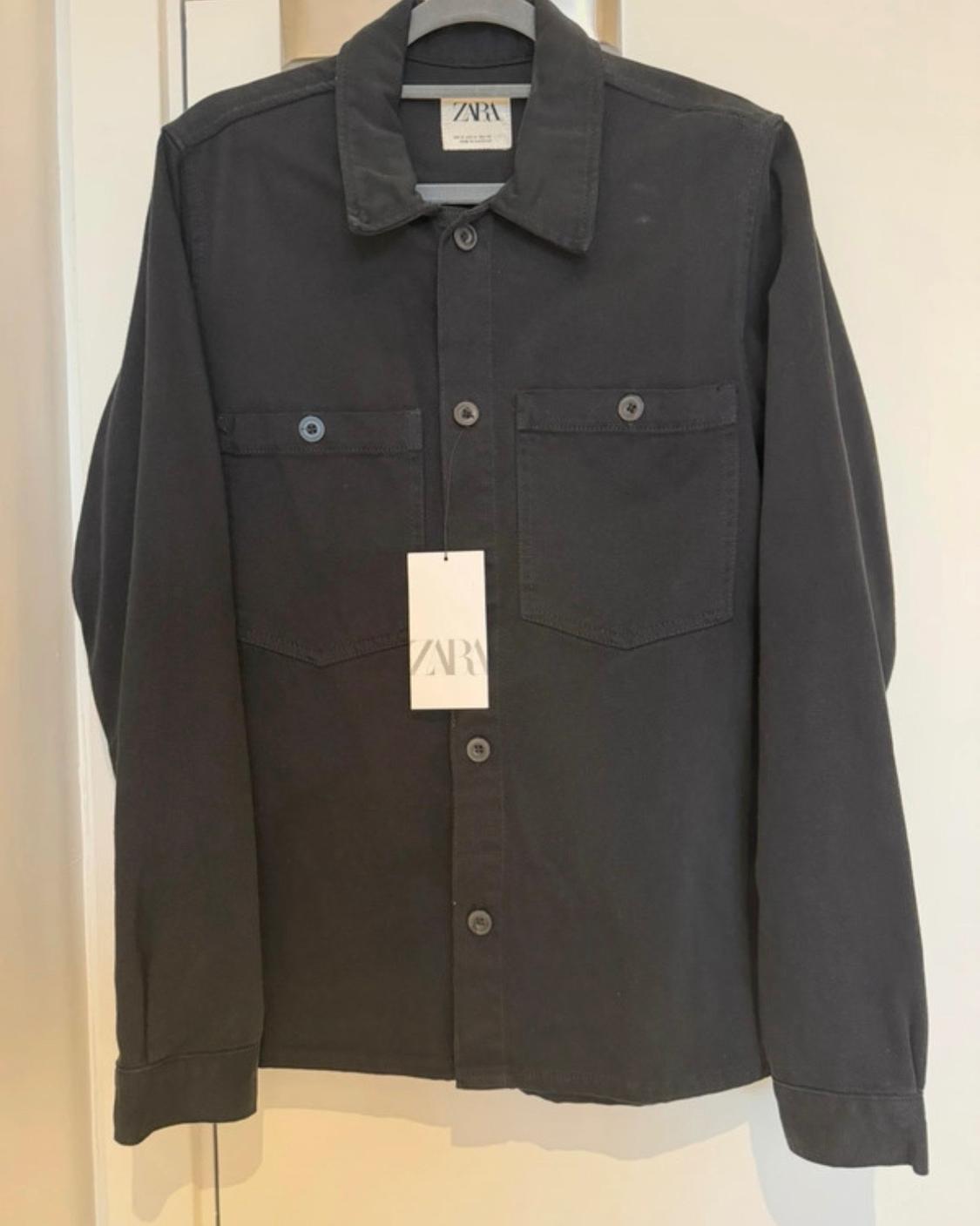 Long-sleeved black shirt hanging on a hanger. Front button fastening, chest pockets and labels with the text ZARA.