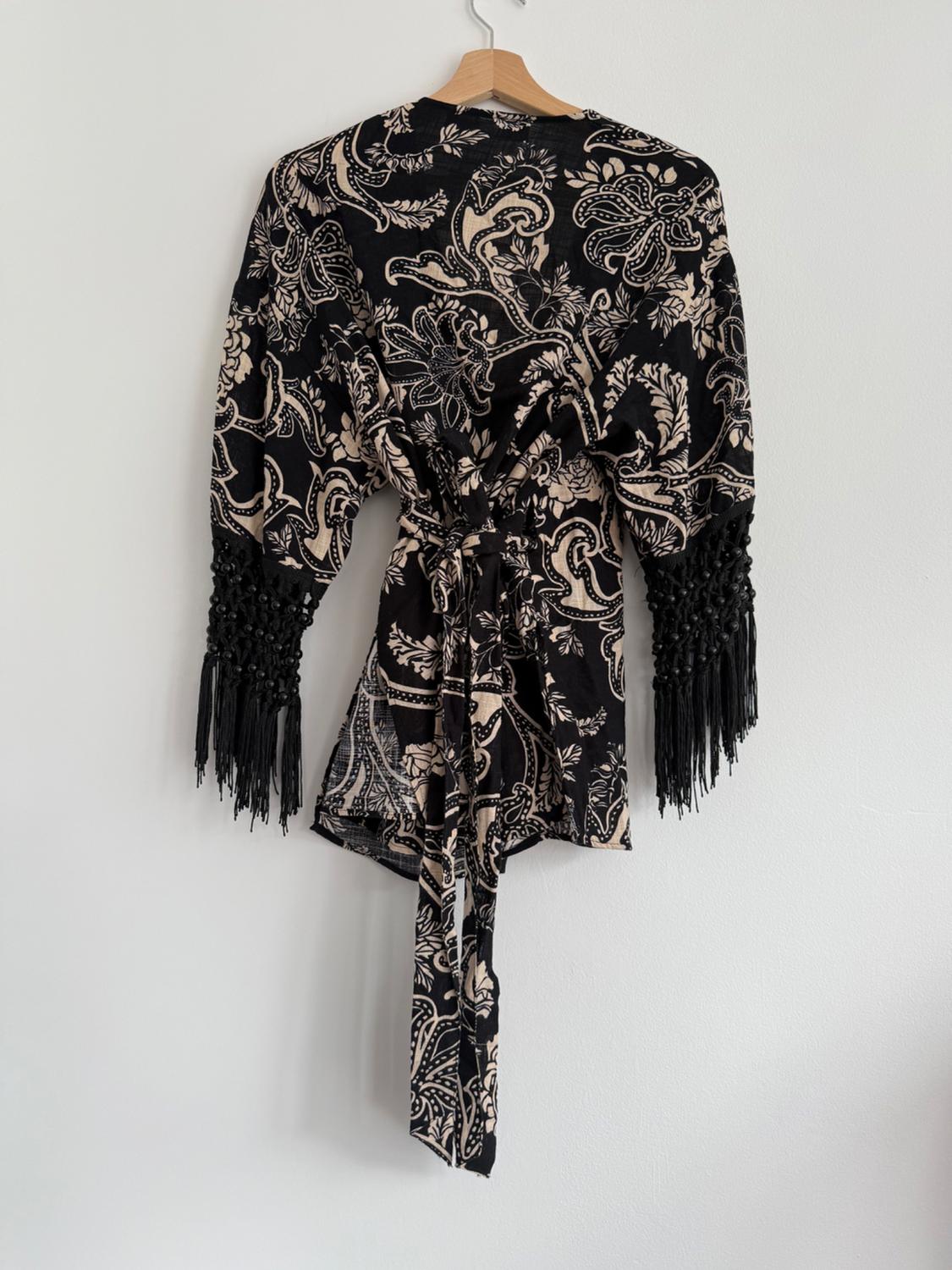 Black kimono with beige floral print, fringed sleeves, hanging on a wooden hanger against a white wall.