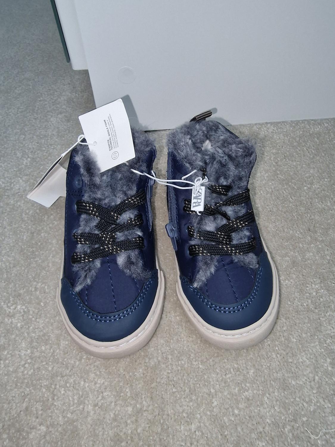 Blue trainers with grey plush lining and black laces on a beige carpet. White tags hanging from the shoes.
