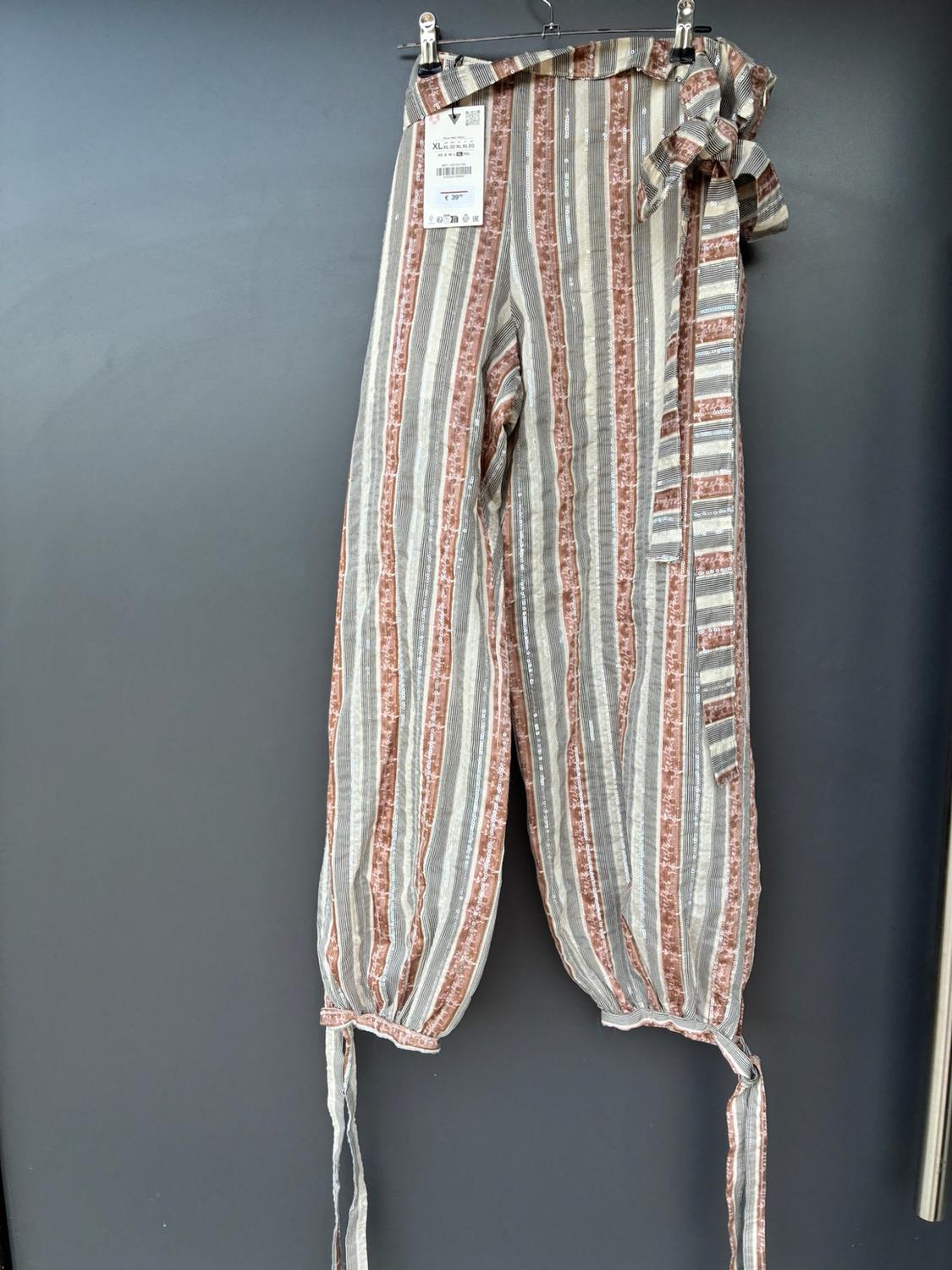 Flowing striped trousers with a tie at the waist and ankles, hanging on a hanger with a label against a grey background.