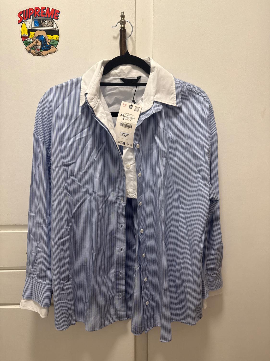 Blue and white striped shirt with a white collar hanging on a hanger. Label visible. "SUPREME" sticker on the wall.