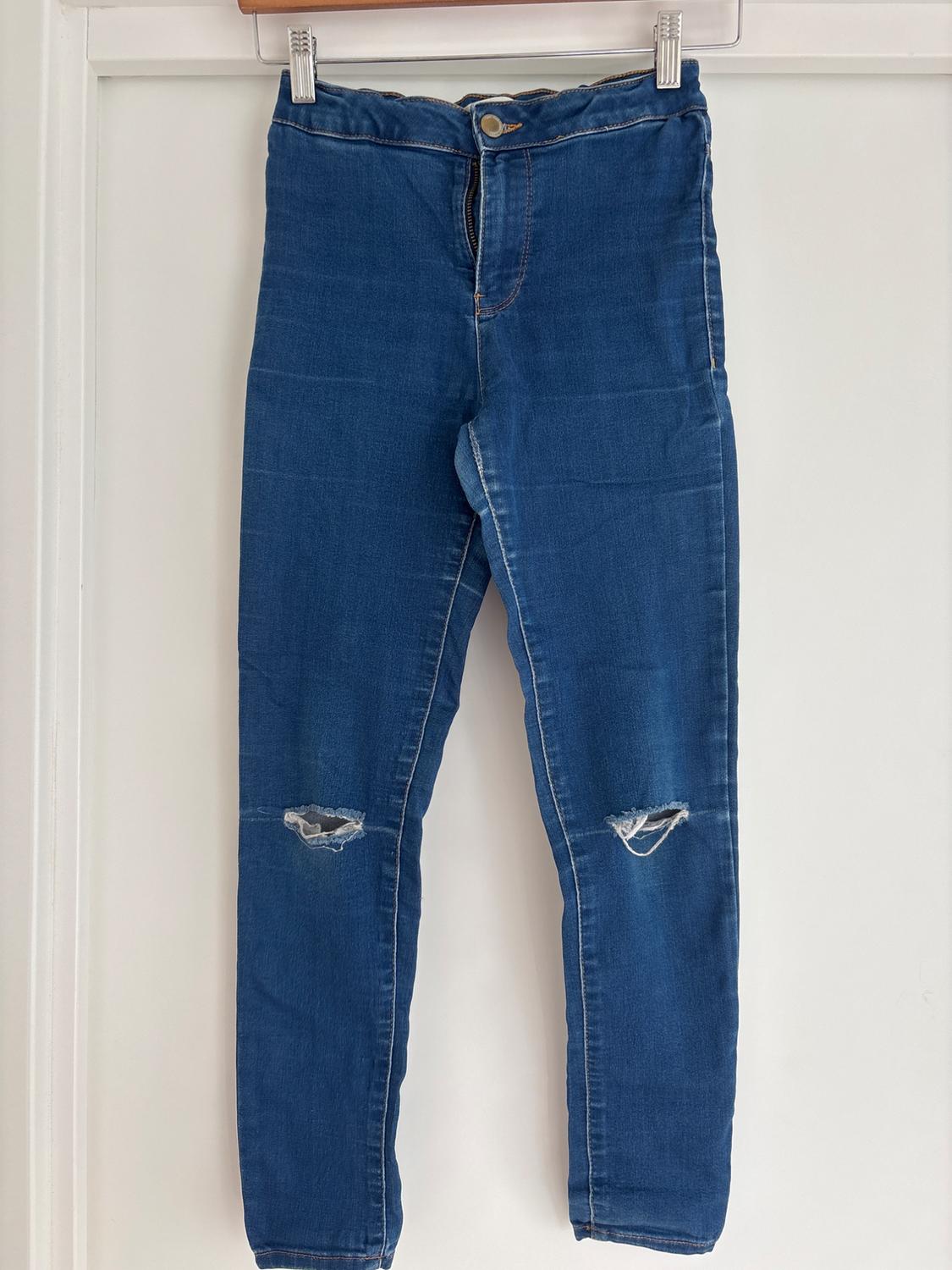 Blue denim jeans hanging on a white hanger, with rips at the knees, against a white background.