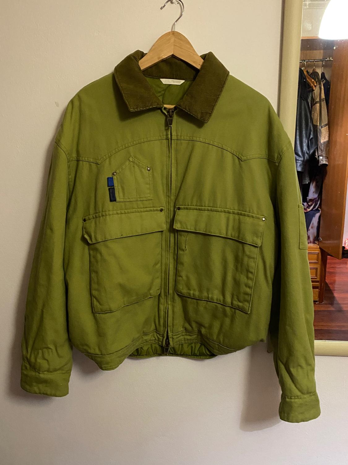 Olive green jacket with a brown corduroy collar, large pockets, and a front zip, hanging on a wooden hanger.