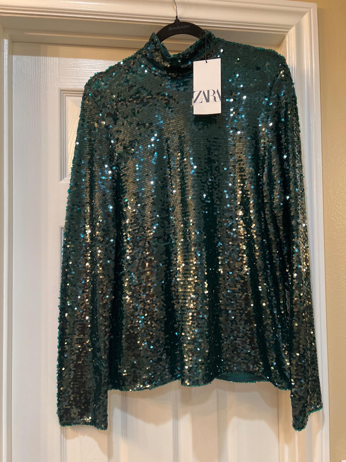 Green sequin jumper hanging on a black hanger with a Zara tag, on a white door.