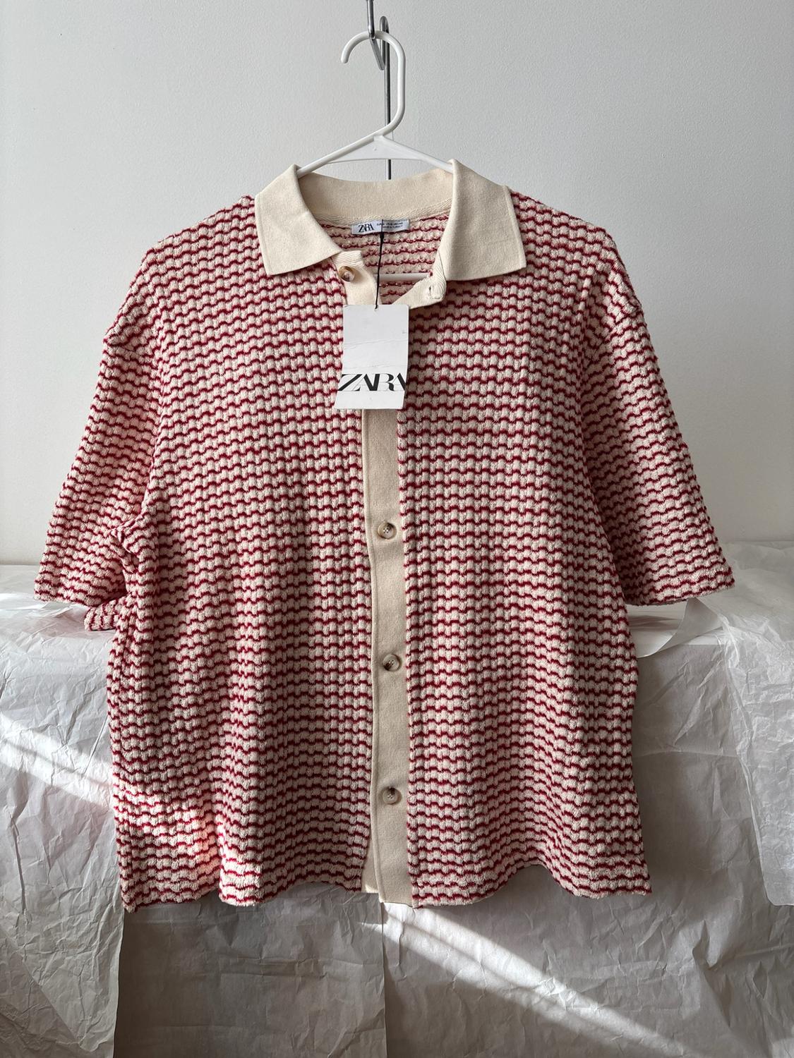 Red and white dot polo with short sleeves, cream collar and buttons. Label with text ZARA. Hung on a hanger over wrinkled white paper.