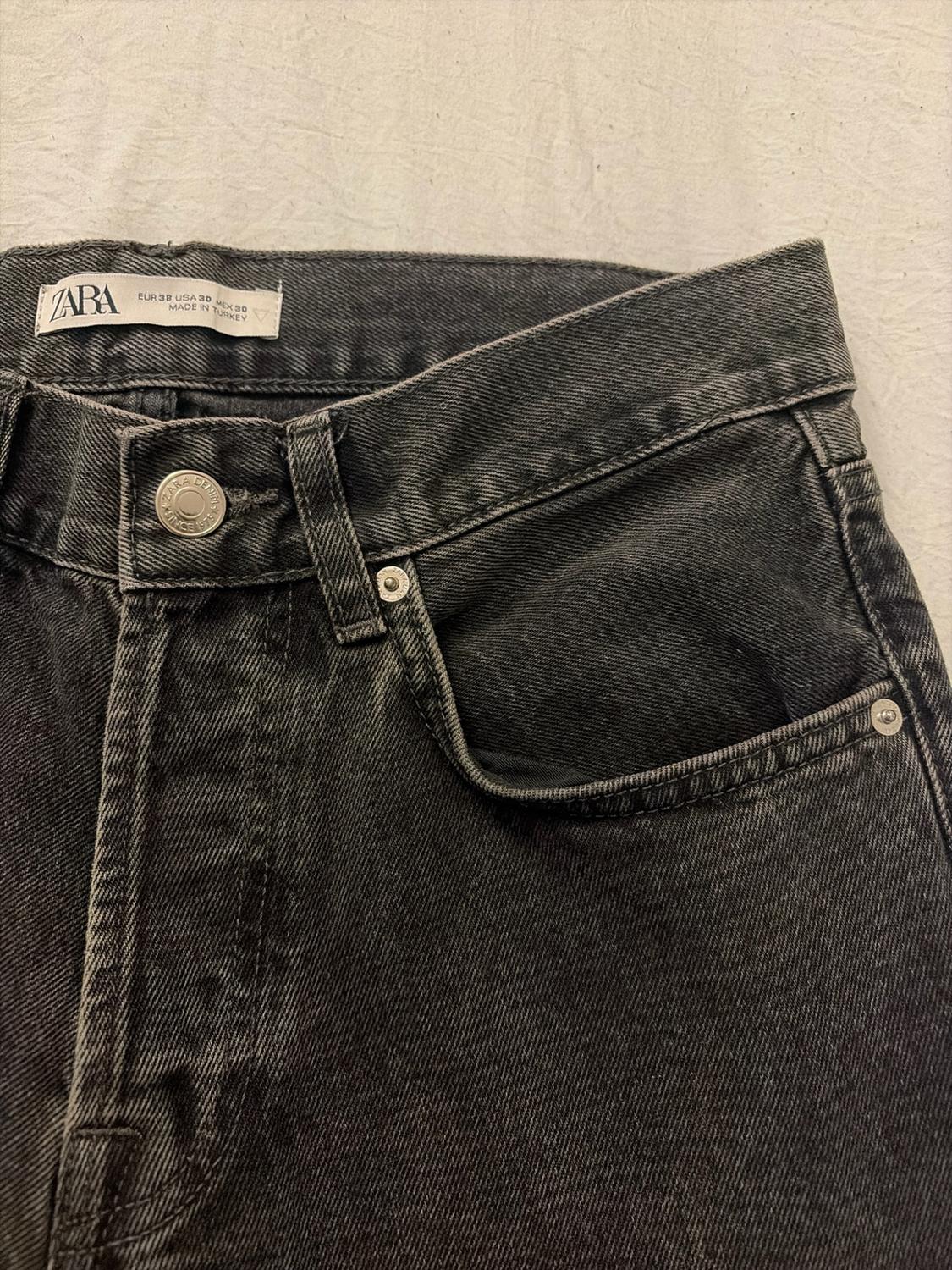 Detail of black jeans on light fabric. Label: ZARA EUR 38 USA 30 MEX 30 MADE IN TURKEY. Button: ZARA DENIM.