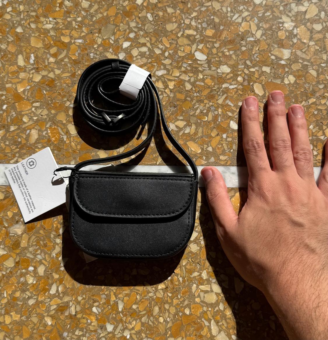 Mini black handbag with flap and rolled strap, placed on a terrazzo floor. A hand positioned next to it emphasises its small dimensions. Label: LEATHER.