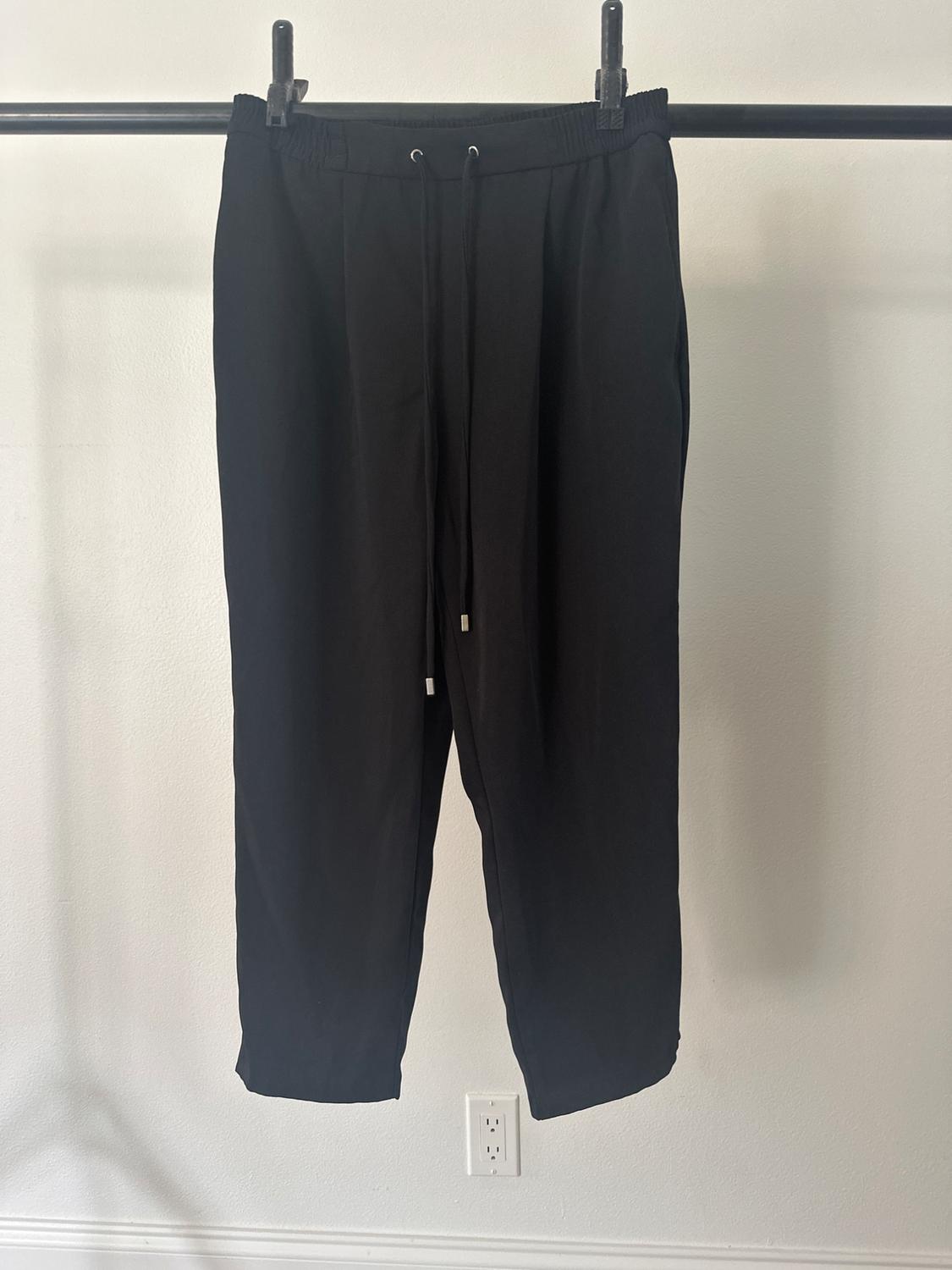 Black trousers hanging on a hanger against a white wall. Elasticated waistband with adjustable drawstrings.