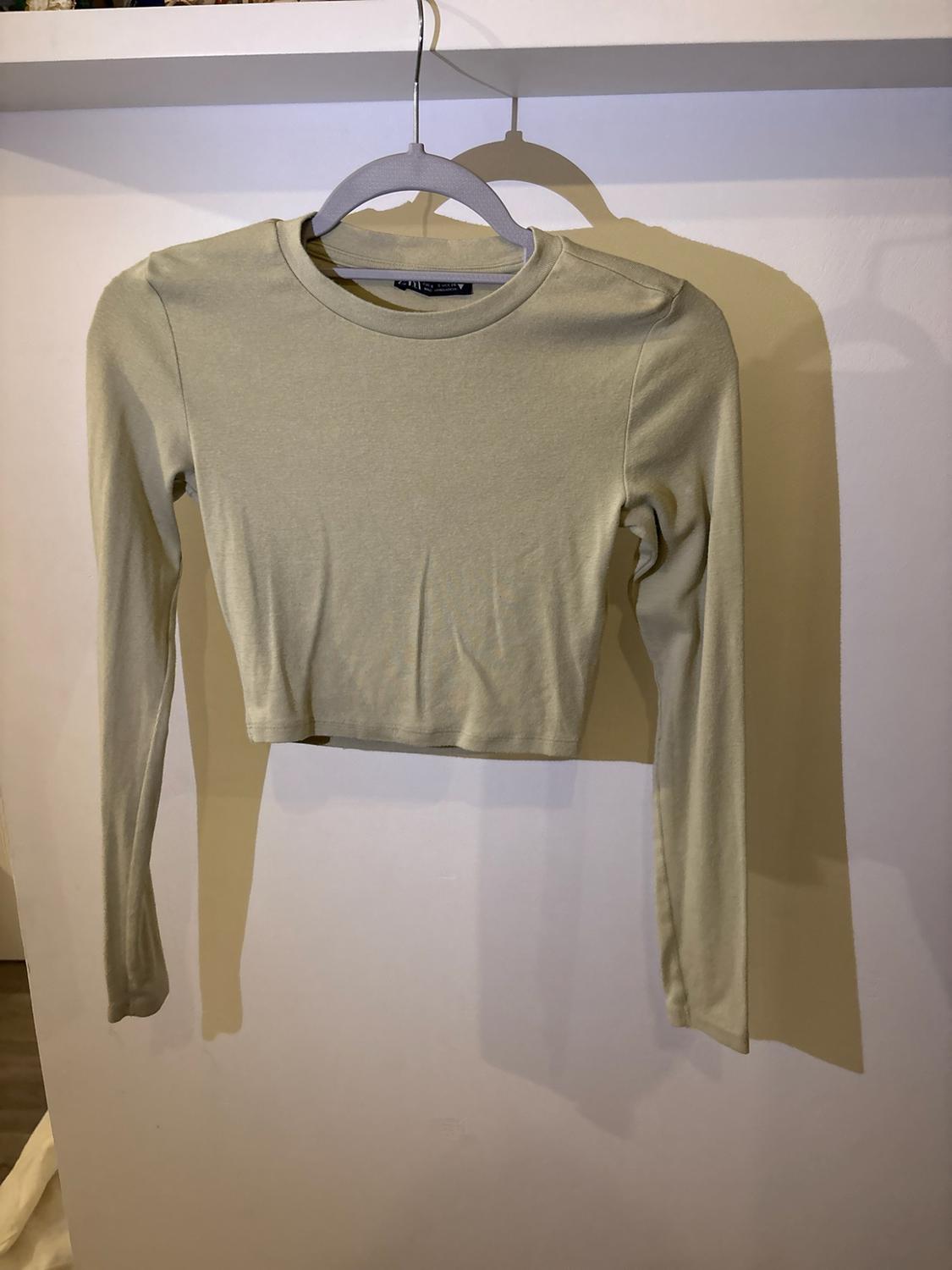 Light green long-sleeved crop top hanging on a grey hanger against a white wall, with cast shadows.