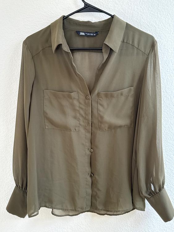 Olive green long-sleeved blouse with buttons and two front pockets, hanging on a black hanger against a white background.