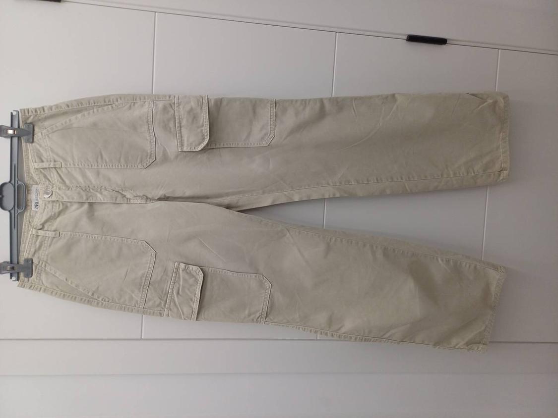 Beige cargo trousers hanging on a grey hanger against a white wall. Large pockets visible on the sides.