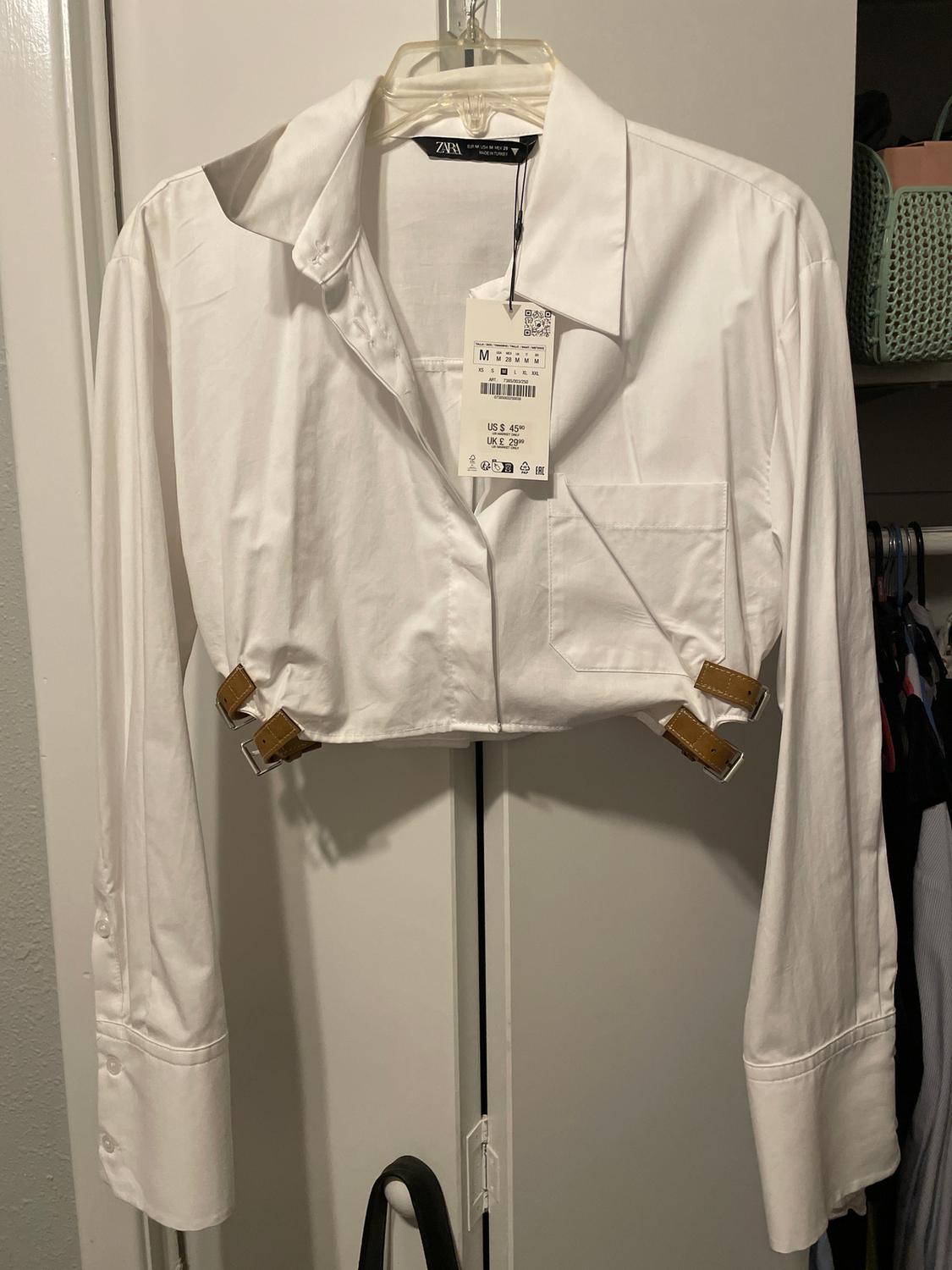 Short white shirt with long sleeves and brown buckles, hanging on a hanger. Visible tag with price and size M.