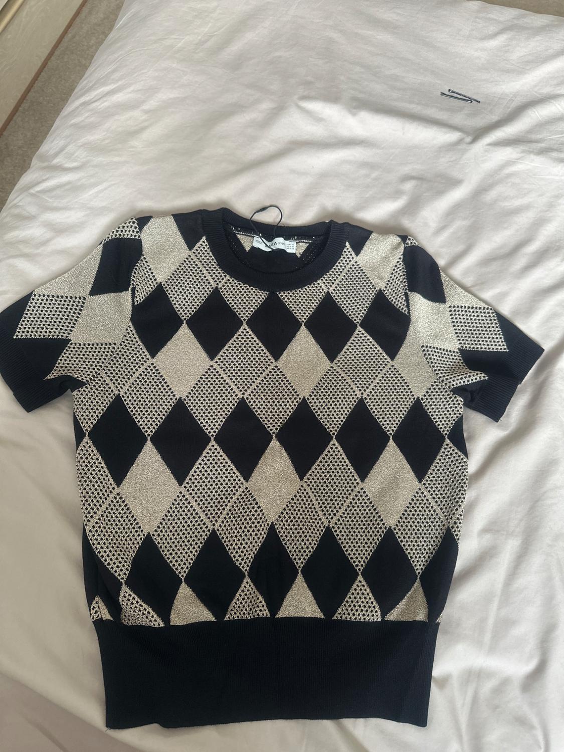 Short-sleeved diamond-knit jumper in black and beige, with a round neck and ribbed trims, laid out on a crumpled white sheet.