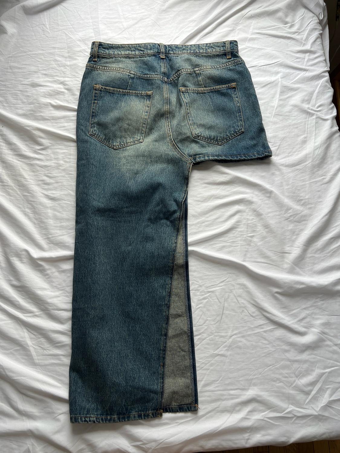 Asymmetric blue jeans with one long leg and one short leg, seen from behind and laid out on a wrinkled white sheet.