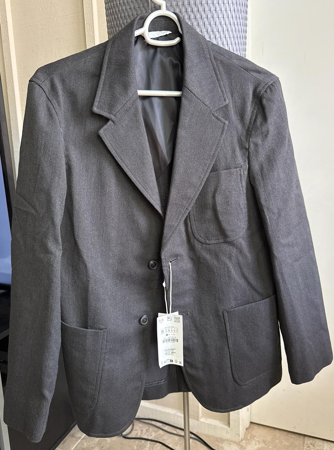 Dark gray blazer on a white hanger indoors. It has two buttons, patch pockets, and a tag with the text S US $109 UK £99.